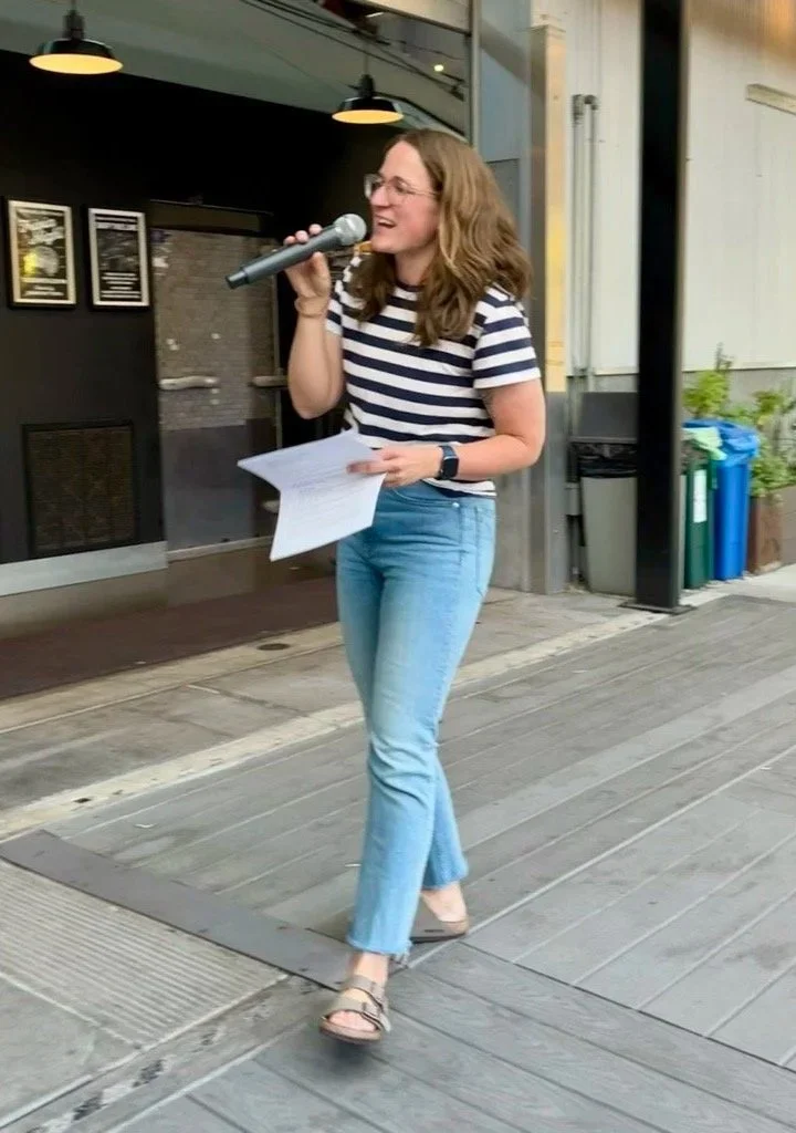A woman with wavy brown hair, glasses, and wearing a striped navy and white t-shirt, light blue jeans, and sandals is smiling while holding a microphone and a piece of paper, standing outdoors on a wooden deck.