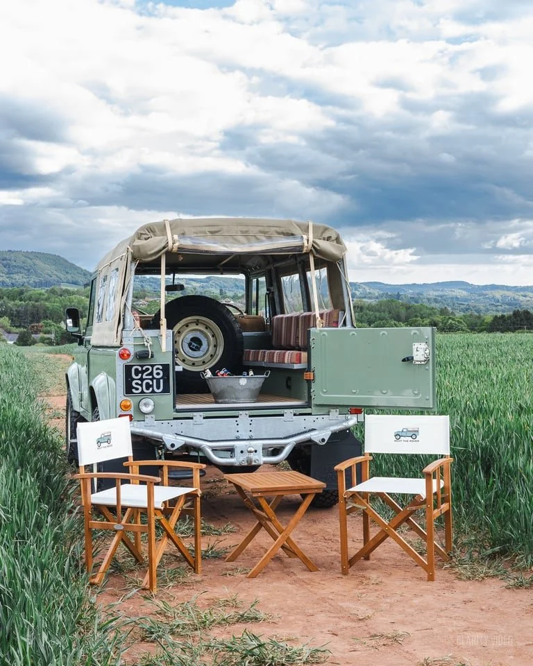 An outdoor scene featuring a vintage Land Rover parked on a dirt path in a grassy field with mountains and cloudy sky in the background. Two wooden chairs and a small wooden table are set up in front of the vehicle, with signs indicating it's a Land 