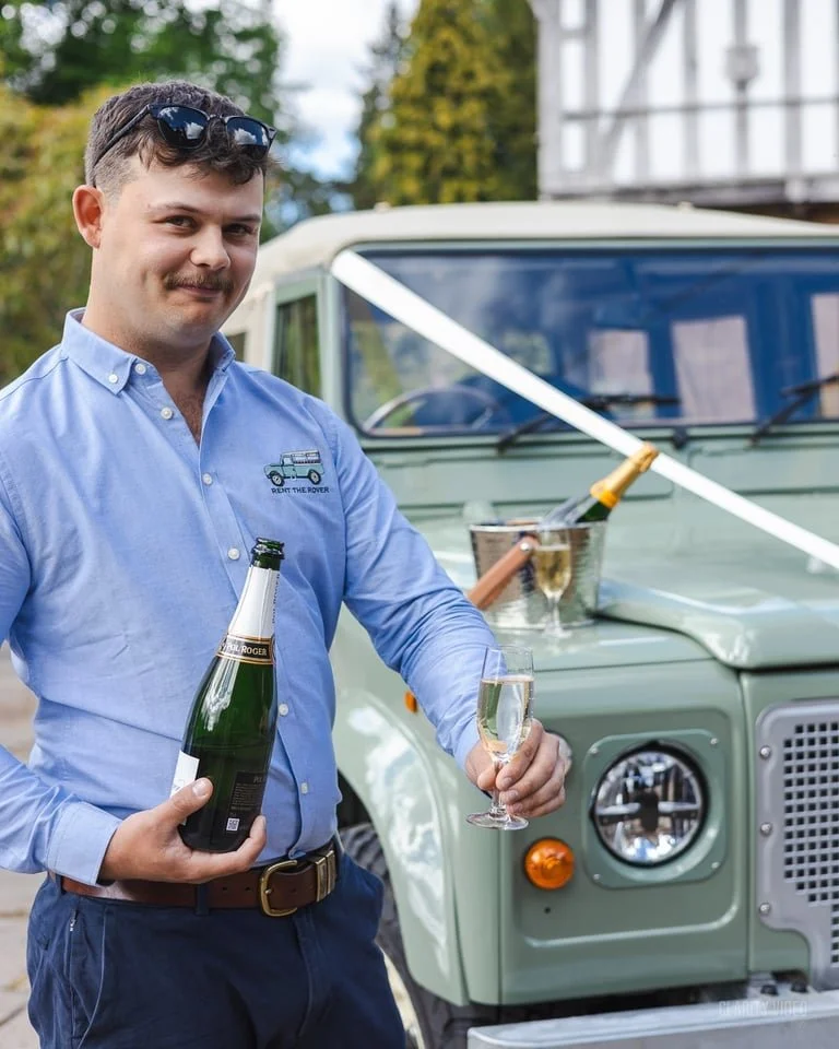 A man with sunglasses on his head, wearing a light blue button-up shirt with a small logo on the chest, stands outdoors holding a bottle and glass of champagne next to a vintage green Land Rover Defender. There is a bottle in an ice bucket on the veh
