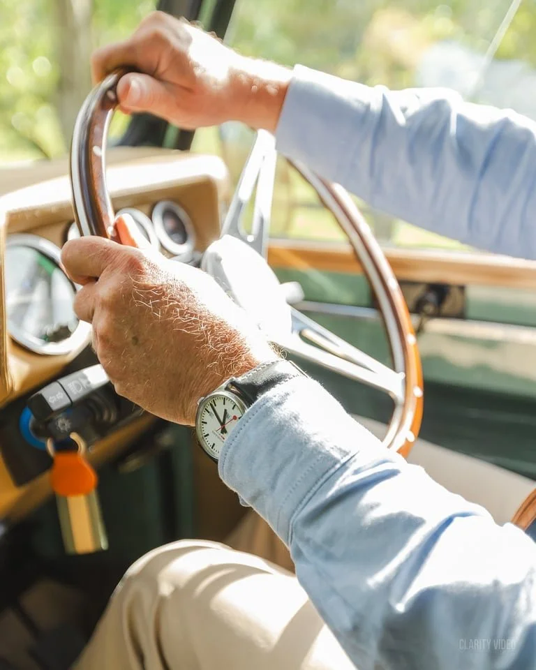 Close-up of a person's hands on a vintage car's steering wheel, with a watch on their left wrist, in a sunlit environment.