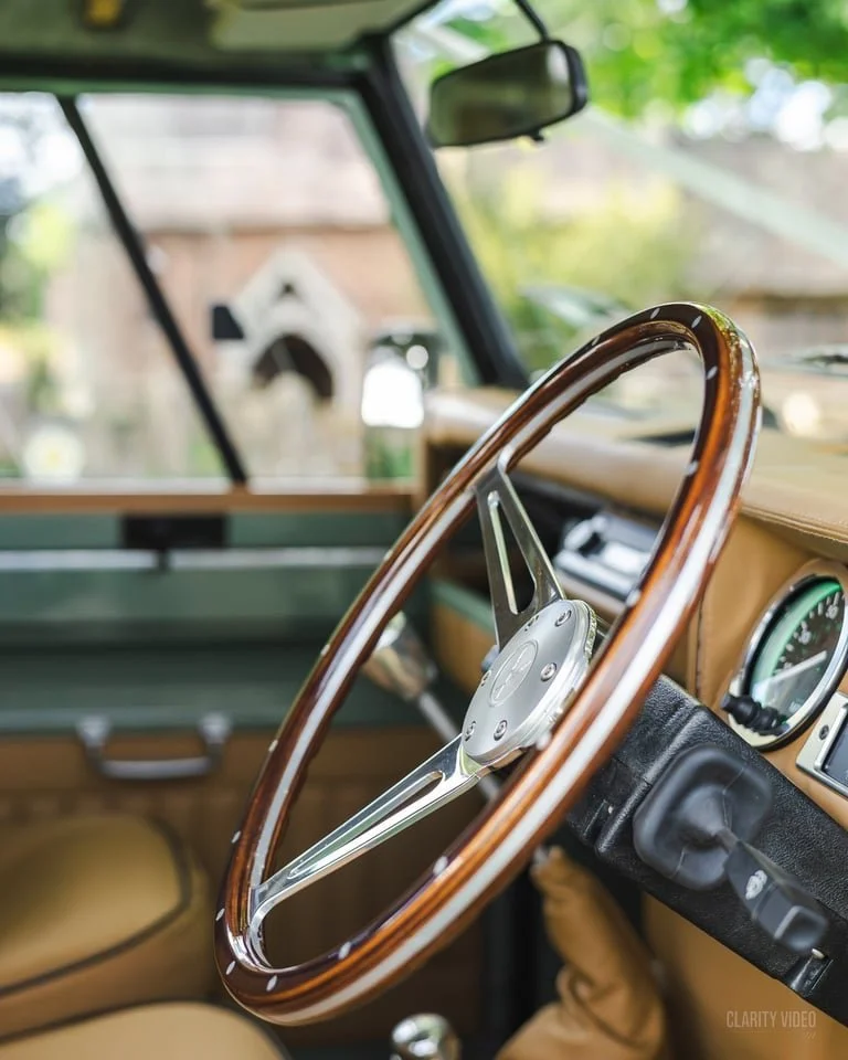 Close-up of a vintage car interior featuring a wooden steering wheel, dashboard with gauges, and a beige and green color scheme.