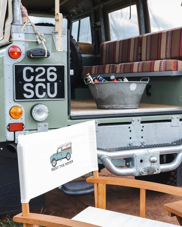Inside a vintage Land Rover with a bench seat and a bucket of bottles, a folding chair with a 'Rent the Rover' sign, and an off-road setting.