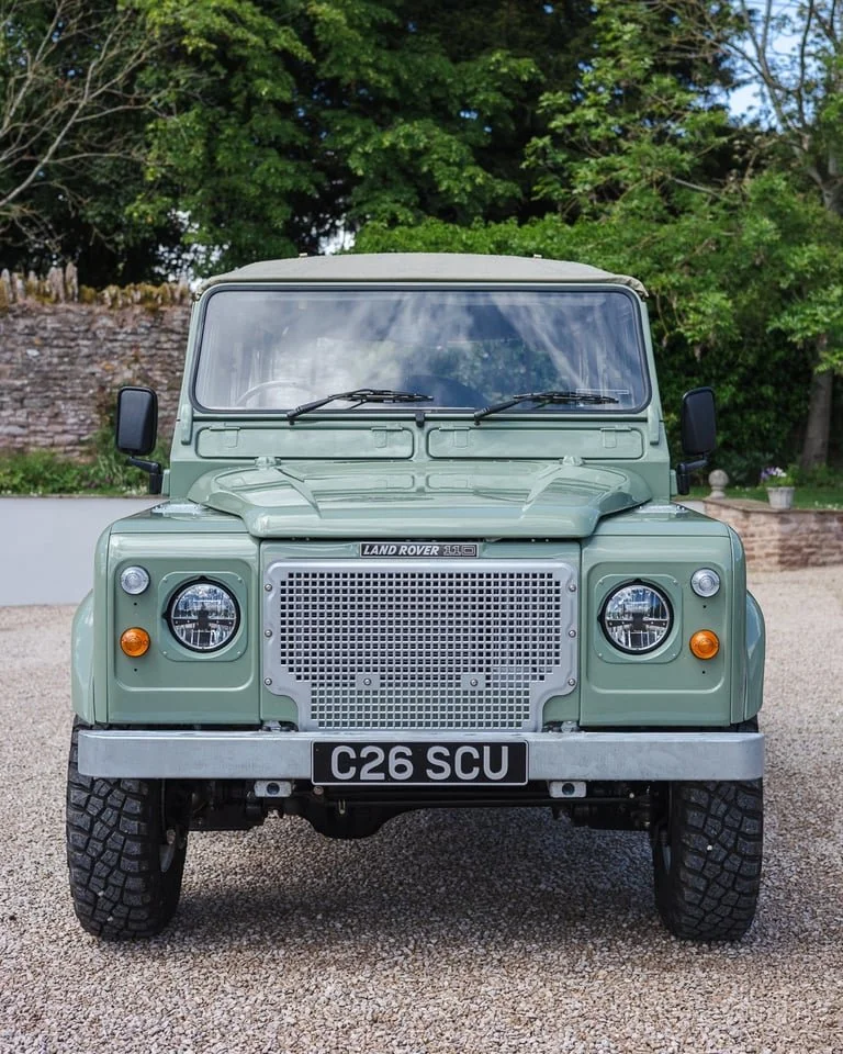 Front view of a mint green Land Rover Defender with black tires, parked on gravel with trees and a stone wall in the background.