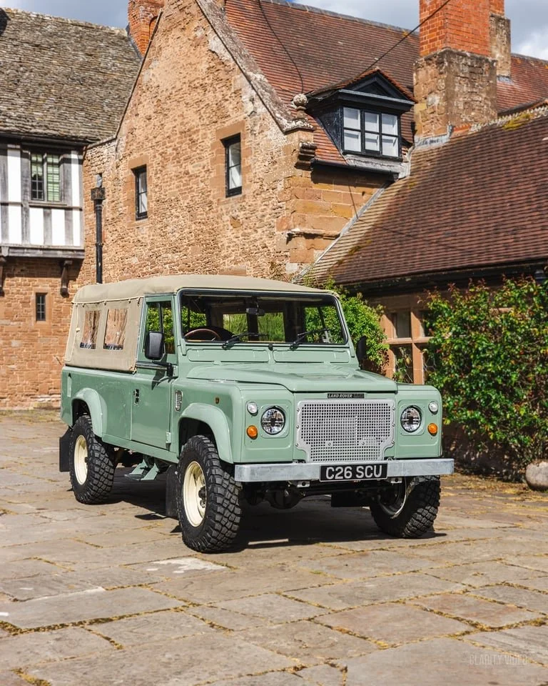 A vintage green Land Rover with a beige canvas top parked on a cobblestone street in front of old brick buildings with steep roofs and chimneys, some greenery on the side.