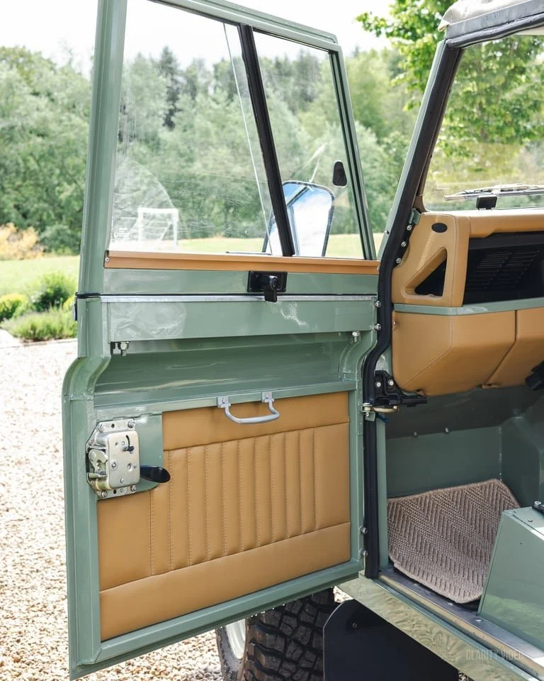 Open vintage vehicle door showing tan interior panel and glass window, with a lush green outdoor scenery in the background.