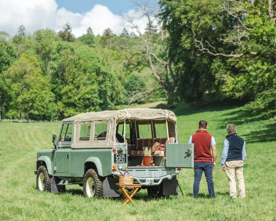 Two men standing near an open-back vintage green Land Rover in a grassy field surrounded by trees.