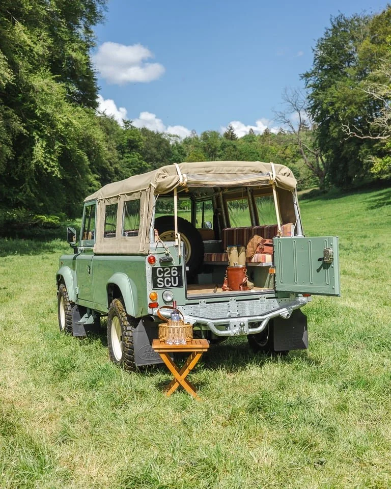A vintage light green off-road vehicle with a beige canvas top parked on a grassy field surrounded by trees, with a small table set up nearby holding bottles and glasses, suggesting a picnic or outdoor gathering.