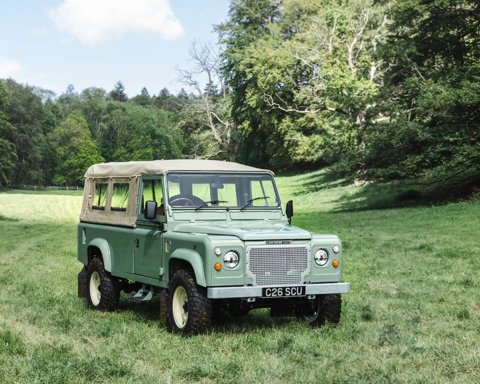 A vintage green Land Rover Defender with a beige canvas top parked on a grassy field surrounded by trees.