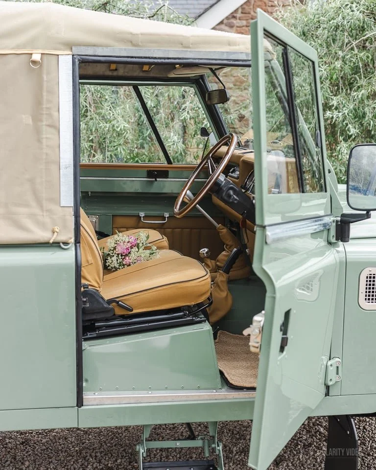 Interior of a vintage mint green Jeep with tan seats, a wooden steering wheel, and a small bouquet of flowers on the seat.