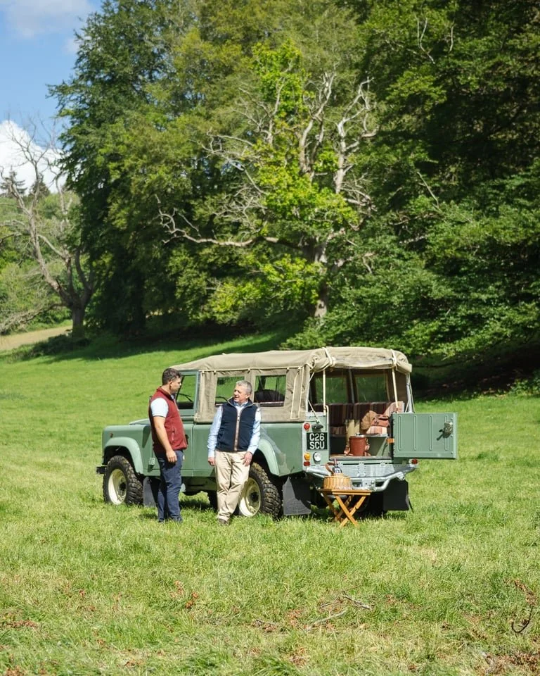 Two men standing next to a vintage green off-road vehicle with a beige canvas top, parked on a grassy field with trees in the background. There is a small wooden table with baskets and supplies near the vehicle.
