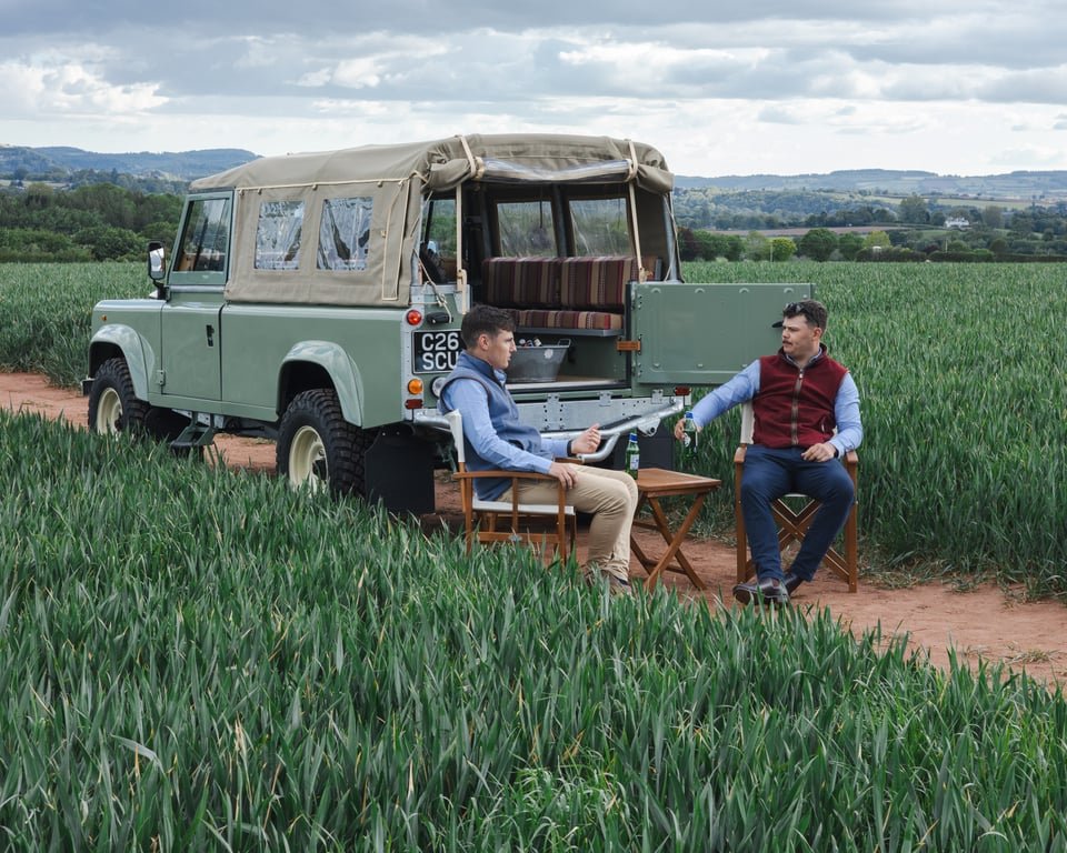 Two men sit on wooden chairs and chat beside a vintage off-road vehicle in a field of green crops, with rolling hills and cloudy skies in the background.