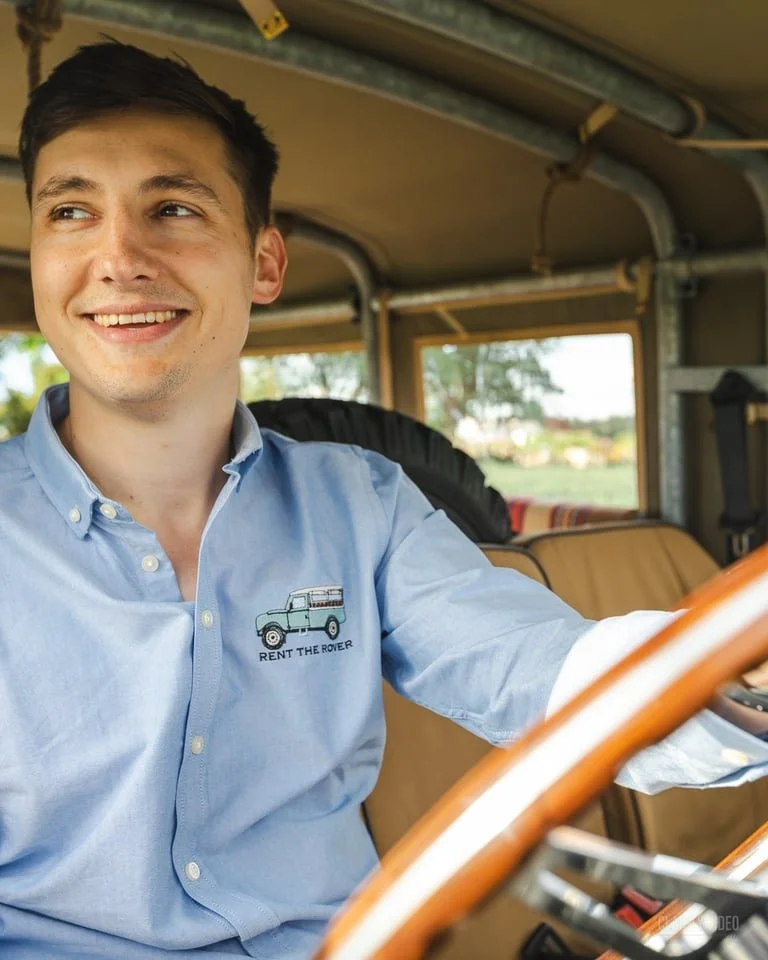 A young man with dark hair and a light blue button-up shirt smiling while sitting at a vintage vehicle's dashboard during daytime, with outdoor scenery visible through the windows.