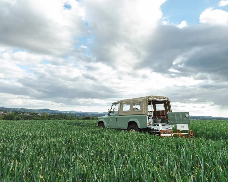 An old green Land Rover with a beige soft top parked in a lush green field of what appears to be wheat or tall grass under a cloudy sky.