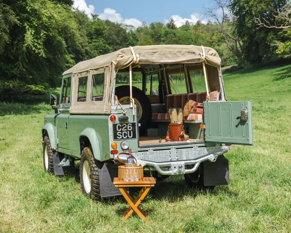 Green vintage off-road vehicle with beige canvas top parked in a grassy field with trees in the background. The back door is open, revealing a striped bench seat and various camping gear. A small folding table with glasses and a wicker basket is set 