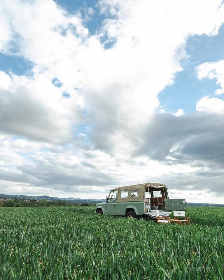 A vintage off-road vehicle with a beige canopy and open rear, parked on a lush green field, under a partly cloudy blue sky.