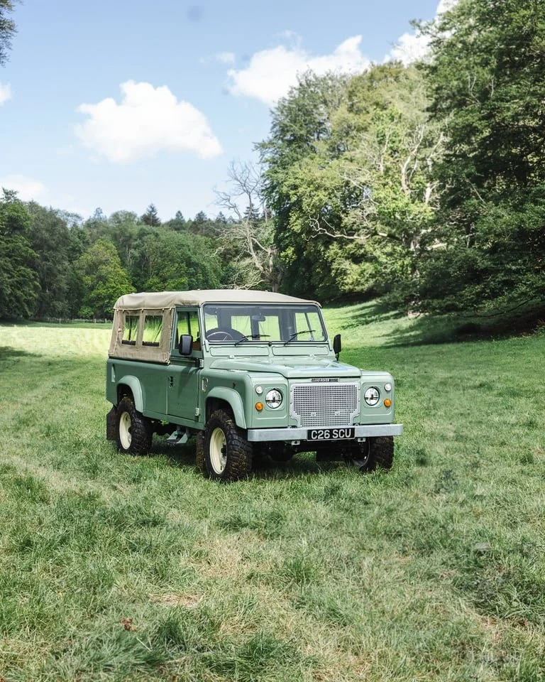 A vintage green Land Rover parked in a lush green field with trees in the background and a partly cloudy sky.
