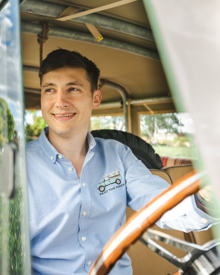 Man sitting inside a vintage vehicle, smiling, wearing a light blue shirt with a 'Rent the Rover' logo.