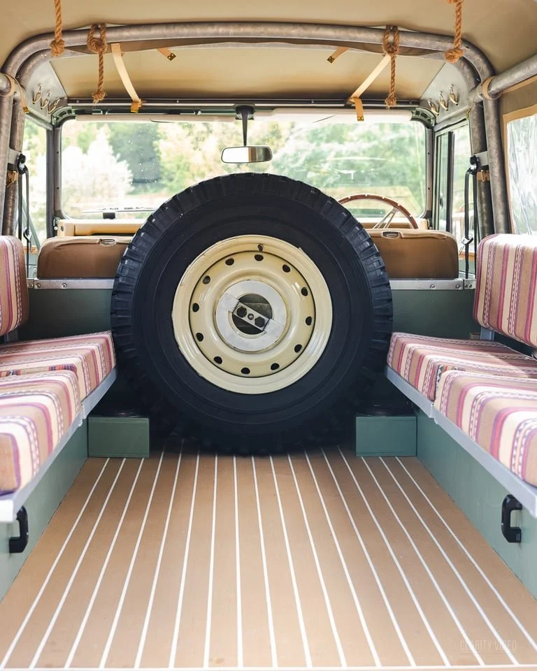 Interior of a vintage off-road vehicle with rear seating, a large spare tire mounted at the back, striped seat cushions, and a wooden floor with white lines.