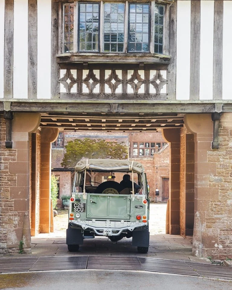 Vintage green military-style vehicle passing through a historic stone archway in a brick building with a wooden balcony above.