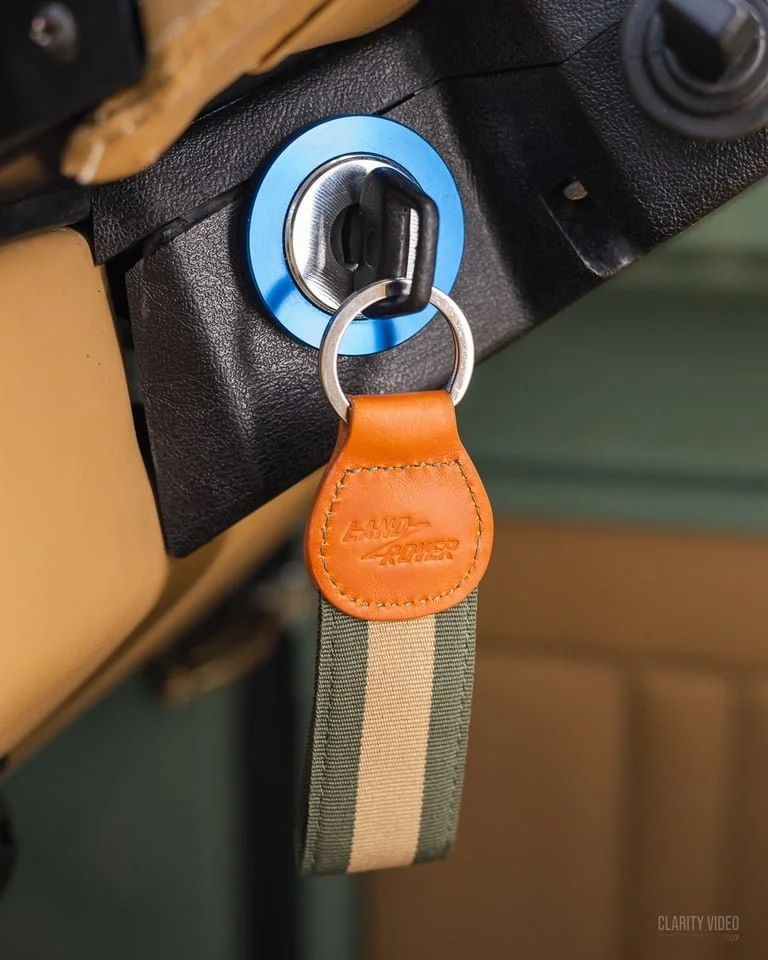 Close-up of a vehicle ignition with a key inserted, attached to a fabric keychain with a leather tab marked 'LANY AIDER'.