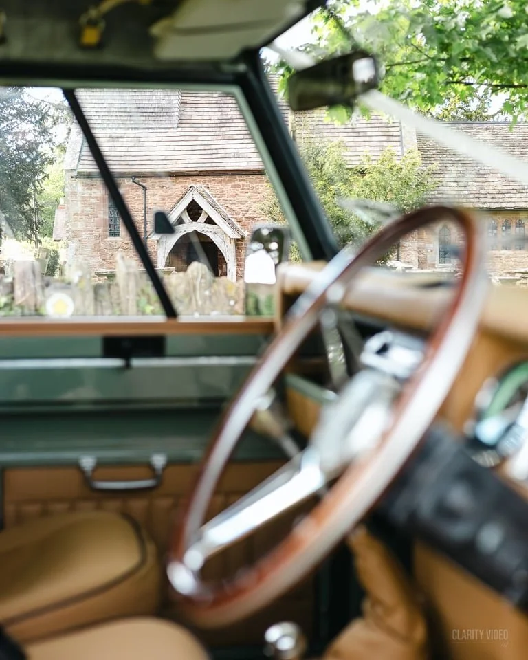View through the windshield of a vintage car showing a brick church with a pointed roof and wooden details, surrounded by greenery.