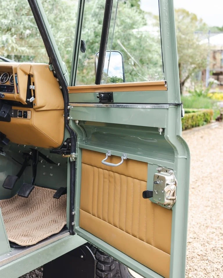Close-up of an open vintage vehicle door with tan interior panels, a handle, and locking mechanisms, with a view of the dashboard and outside greenery.