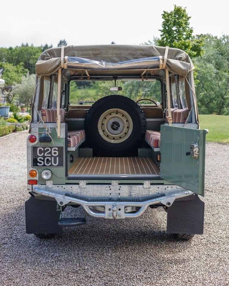 Back of vintage open-top Jeep with spare tire, license plate C26 SCU, and bench seats in a grassy yard with trees.