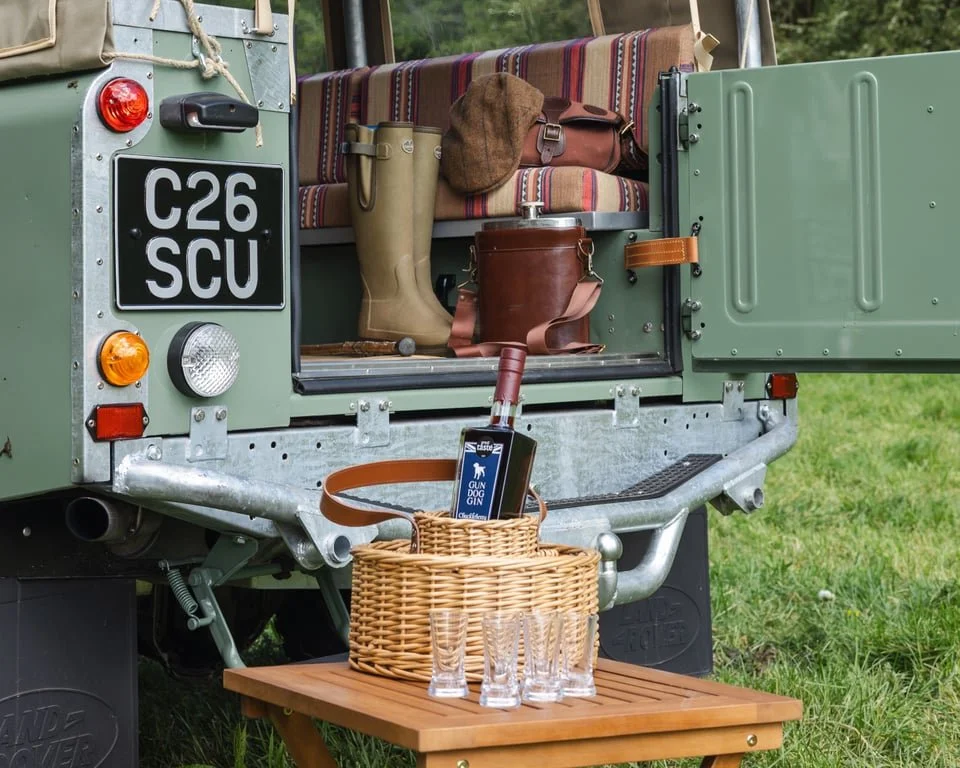 Open back of a vintage green off-road vehicle with camping gear including a blanket, a brown hat, a pair of rubber boots, and a leather bag. A wicker basket on a small wooden table outside the vehicle contains a package of dog treats and a bottle of 