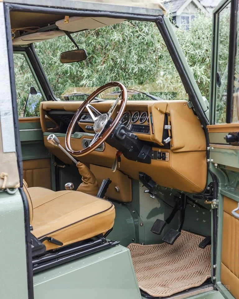 Interior of a vintage military-style vehicle with tan seats, a brown wooden steering wheel, and a dashboard with multiple gauges, surrounded by greenery seen through the windows.