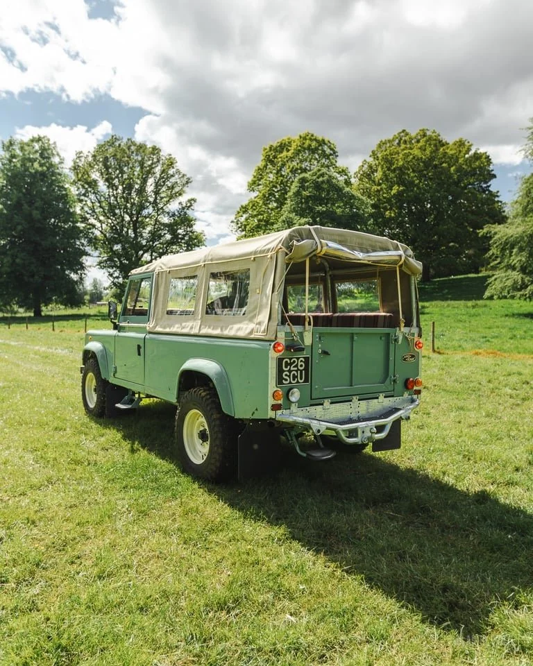 Green vintage off-road vehicle with beige canvas top parked on grass in a park with trees and partly cloudy sky.