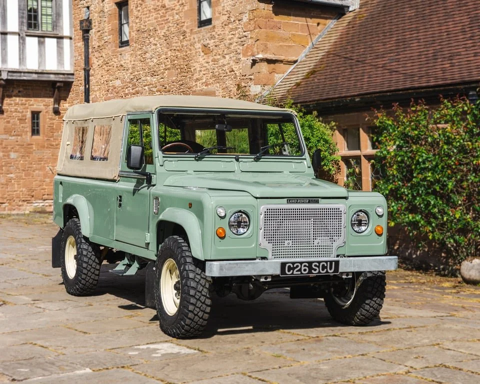 A vintage light green Land Rover Series vehicle with a beige soft top, parked on stone pavement in front of an old brick building with green bushes.