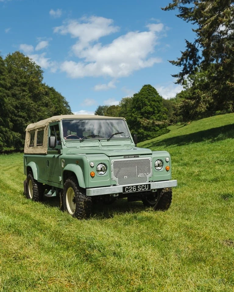 A vintage green Land Rover Defender parked on a grassy field with trees in the background under a partly cloudy sky.