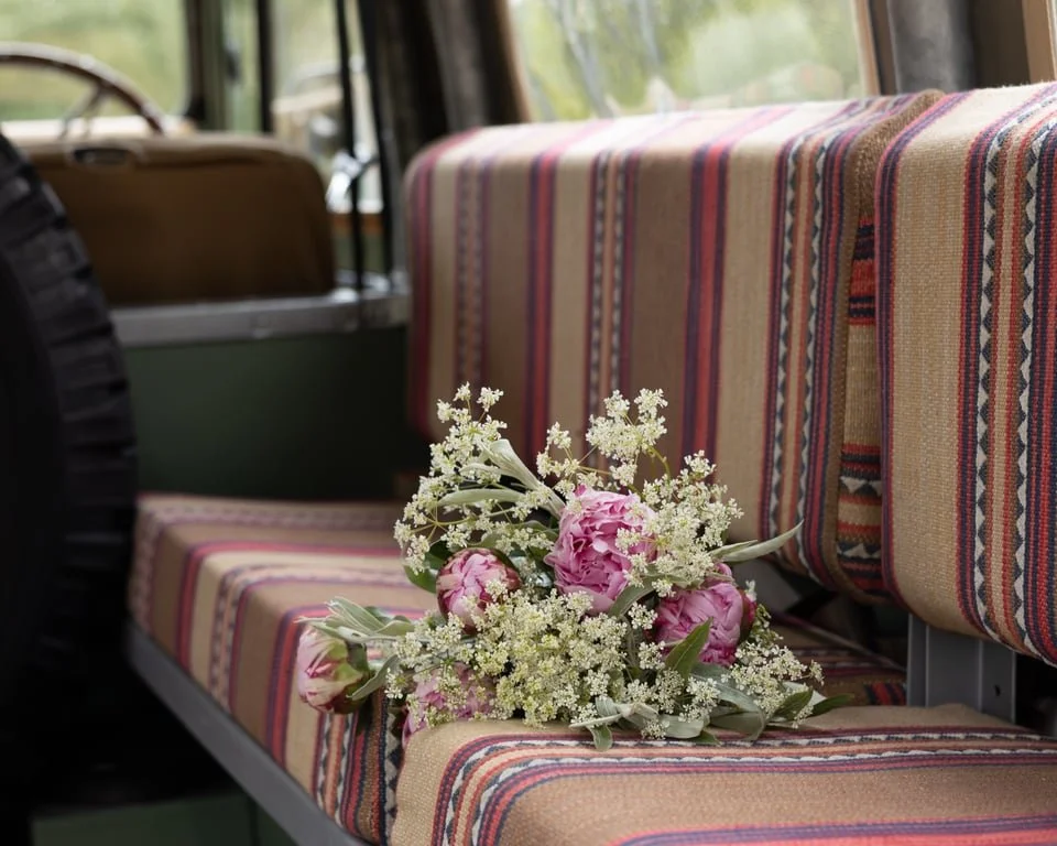 A bouquet of pink peonies and white wildflowers on a striped, vintage van seat with a plaid pattern and a spare tire in the background.