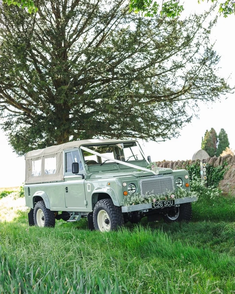 A light green vintage Land Rover Defender with a beige fabric top, decorated with flowers, parked on a grassy field under a large tree.