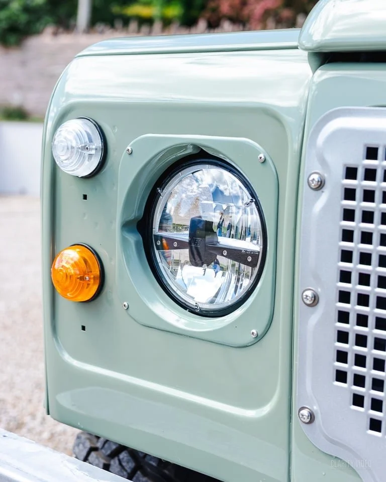 Close-up of a vintage off-road vehicle's front with a large round headlight, smaller clear and amber turn signals, and a metal vent panel.
