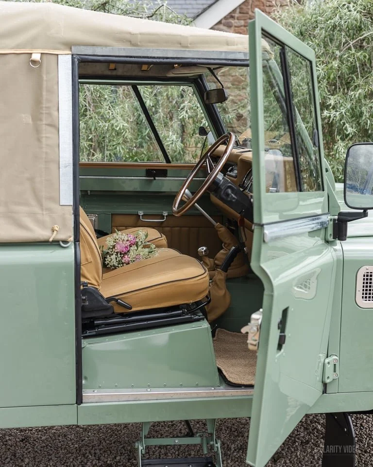 The interior of a vintage green Jeep with a beige fabric covered top, tan leather seats, a bouquet of flowers on the driver's seat, and the driver’s side door open, revealing a simple dashboard and steering wheel.