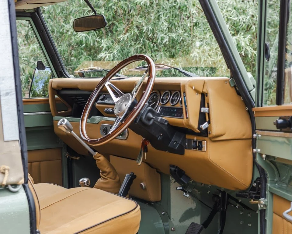 Interior of a vintage off-road vehicle with a tan dashboard, steering wheel, gear shift, and seats, and a green exterior visible through the windows.
