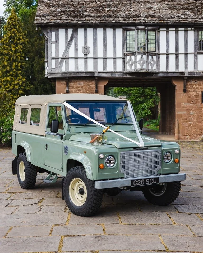 A vintage Land Rover vehicle decorated with a ribbon and a bow on the front, parked on a stone paved area in front of a historic building with half-timbered and brick architecture.