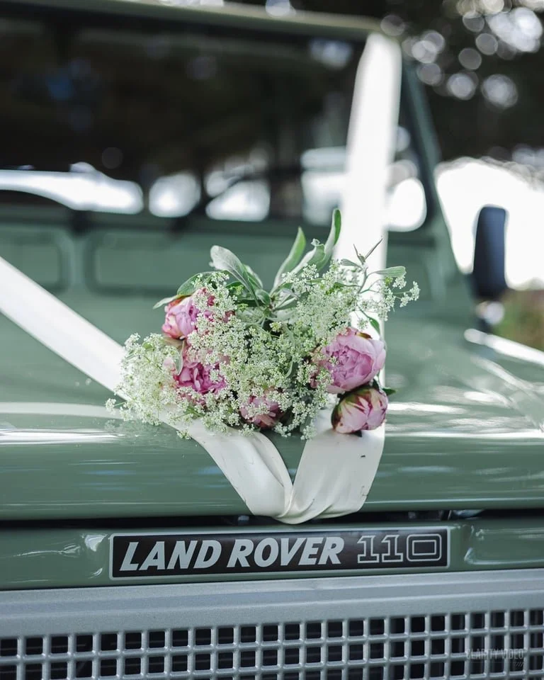 A vintage green Land Rover 110 decorated with a floral arrangement of pink peonies, white baby's breath, and greenery, secured with a white ribbon.