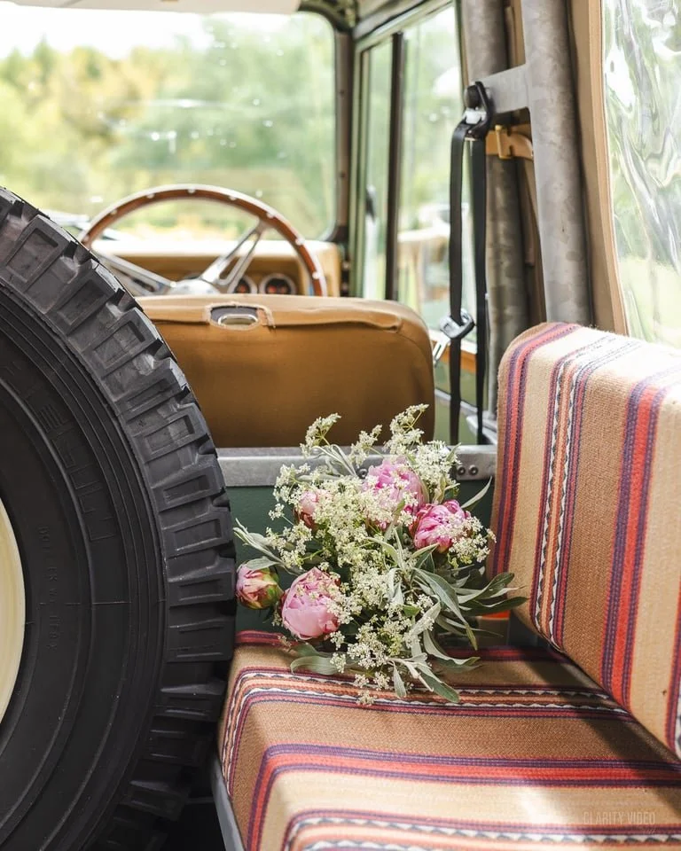 Inside a vintage vehicle, showing a floral bouquet with pink peonies and white filler flowers on a striped upholstered seat with orange, red, purple, and cream colors.