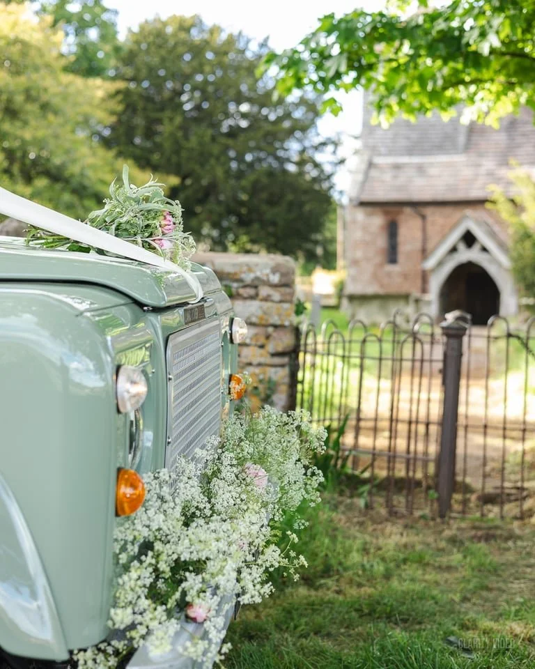 Vintage green car decorated with white and pink floral arrangements, parked outdoors near a stone wall and a small wooden fence, with greenery and a small stone building in the background.