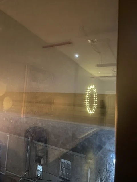 The photo shows a reflection of a room with a circular LED wall light, a map, and some ceiling fixtures, seen through a glass window or door at night.