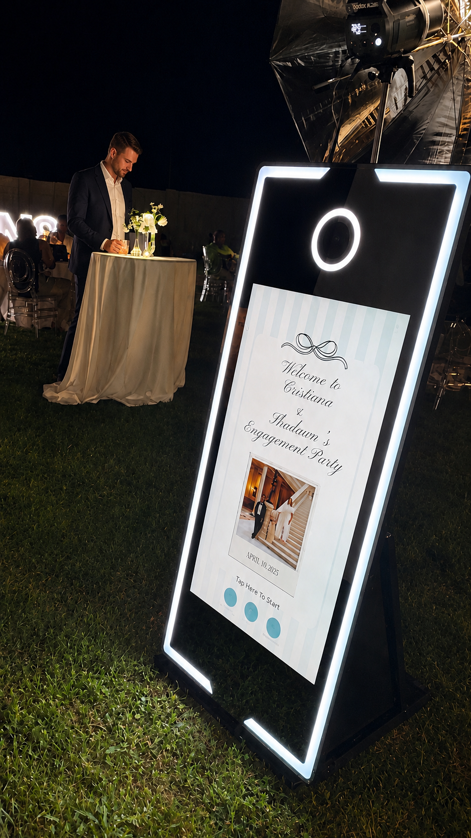 A large illuminated sign at an outdoor engagement party displays a welcome message for Cristiana and dawn, with a photo of a couple on a staircase, the date April 10, 2025, and a prompt to tap to start. In the background, a man in a suit is writing at a tall table with floral arrangements, set up on a grassy area during nighttime.