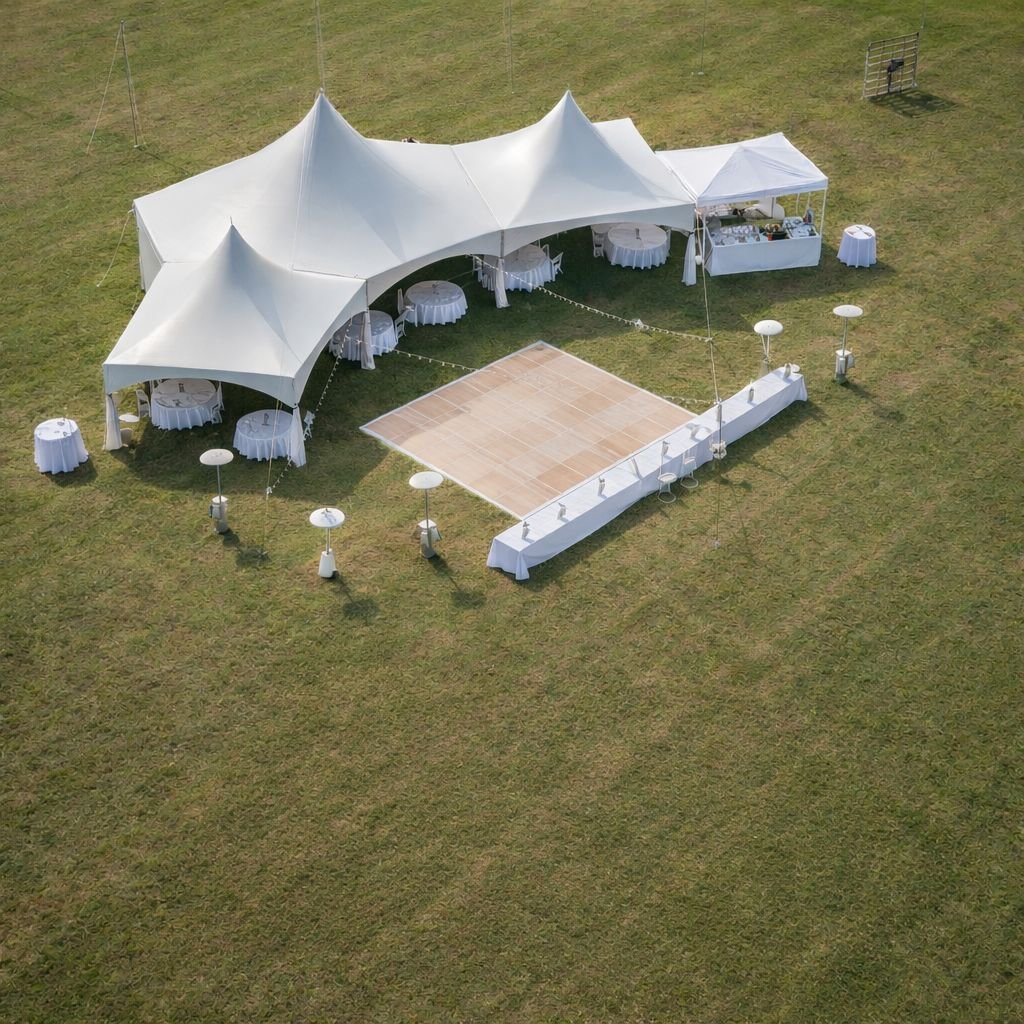 An outdoor event setup with a large white tent, round dining tables with white tablecloths, and a small dance floor area made of wooden tiles, set on a grassy field.