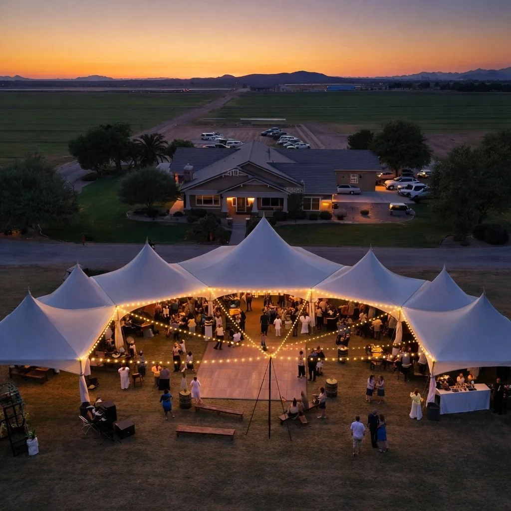 Outdoor wedding reception under a large white tent with string lights, guests mingling, and a backdrop of a rural house and fields at sunset.