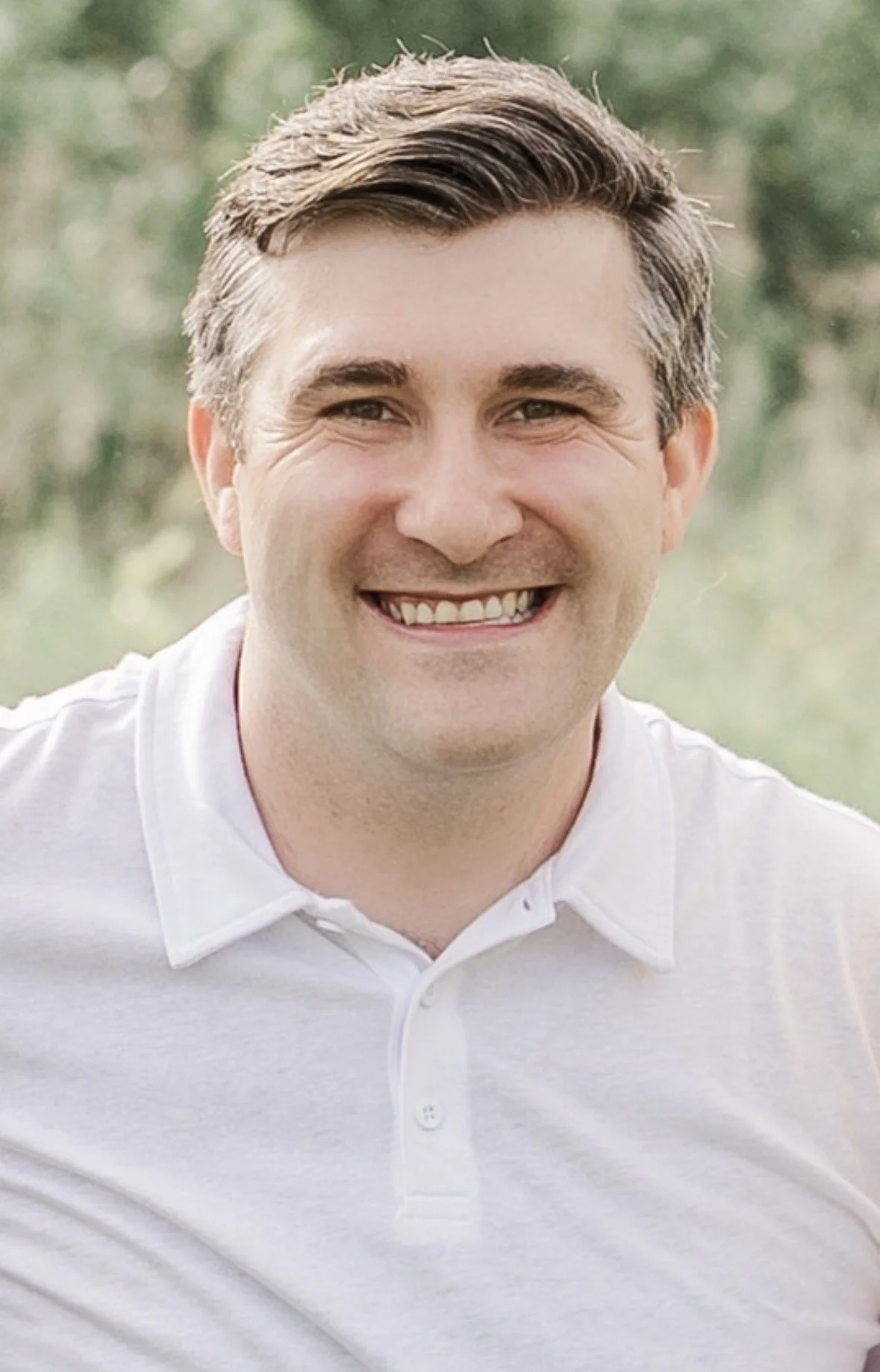 Close-up of a smiling man with short brown hair wearing a white polo shirt, outdoors with blurred green background.