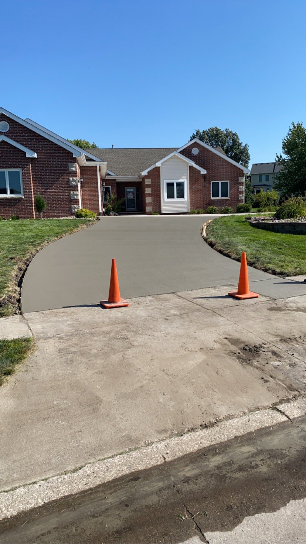 Newly poured concrete driveway with orange traffic cones, leading to a brick house with a white front porch, under a clear blue sky.