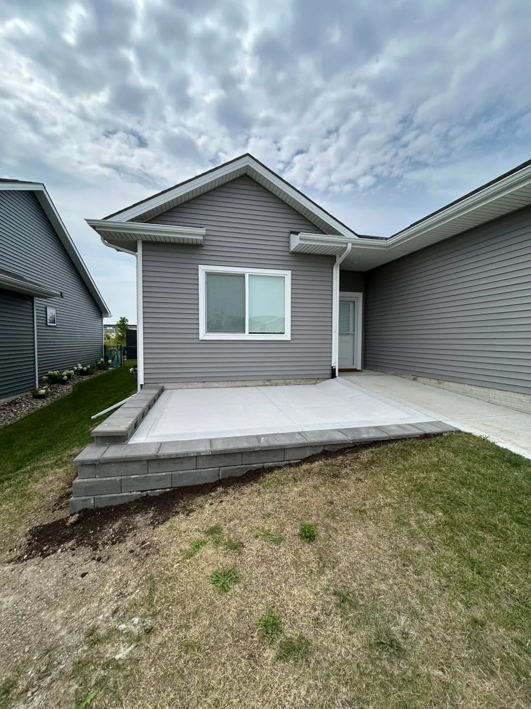 Newly constructed backyard patio with concrete slab and stone border, adjacent to a gray house with vinyl siding and a white-framed window, under a partly cloudy sky.