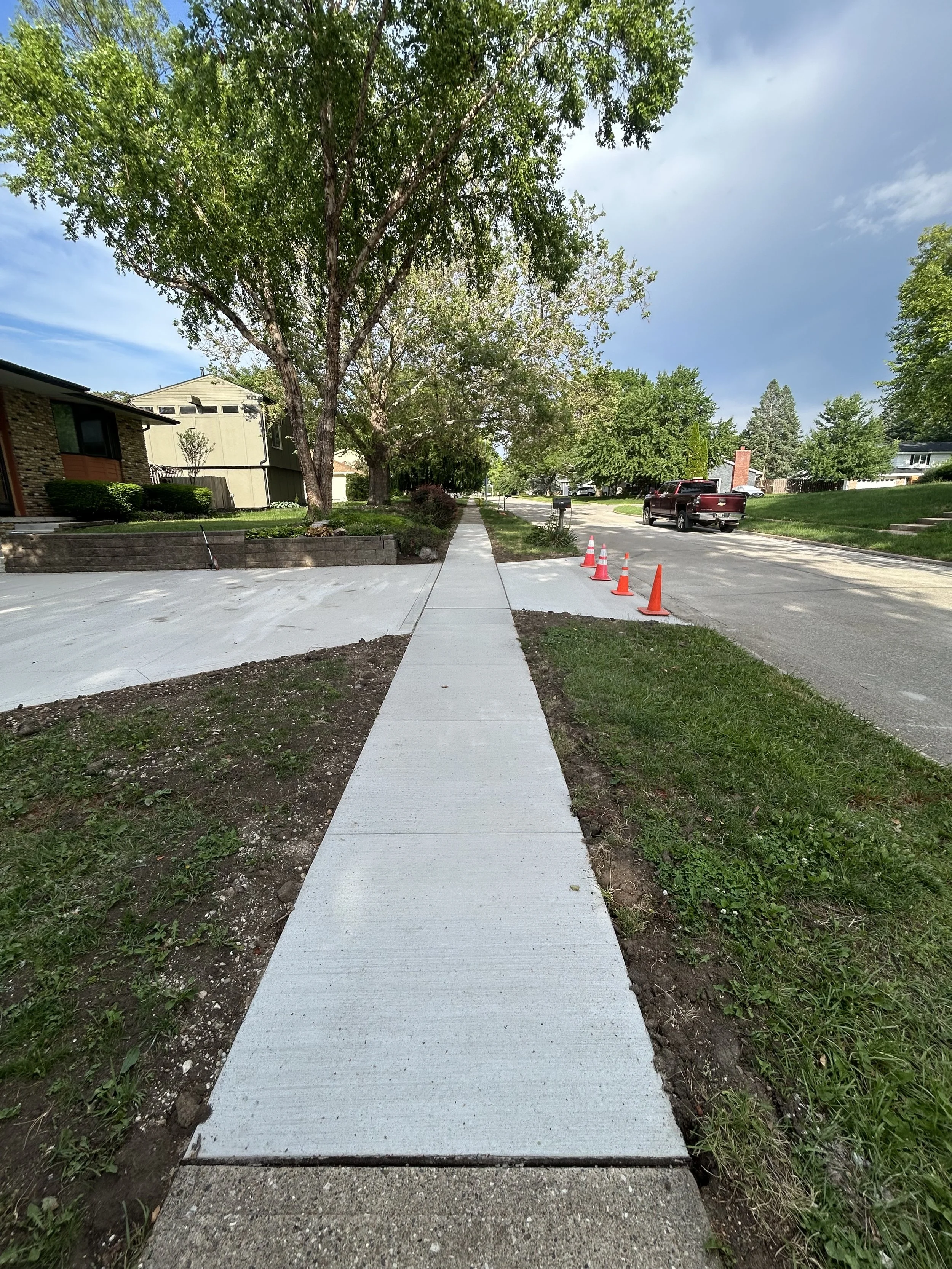 Newly poured concrete sidewalk with a smooth surface and a curb at the end, bordered by grass and soil, with orange traffic cones nearby and a suburban street with parked cars and houses in the background.
