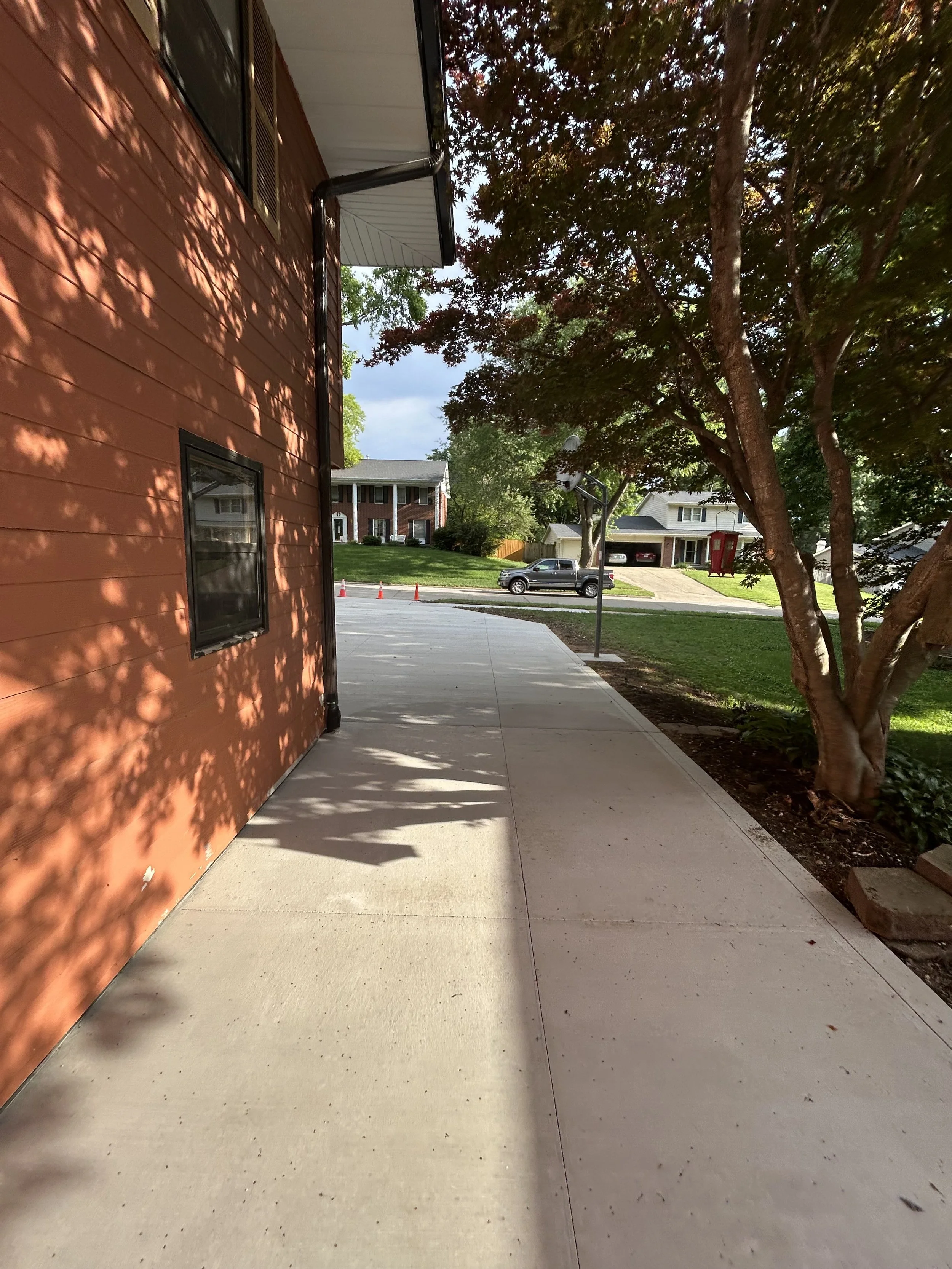 Sidewalk next to a red house with a window, trees casting shadows, and a street with parked cars and houses in the background.
